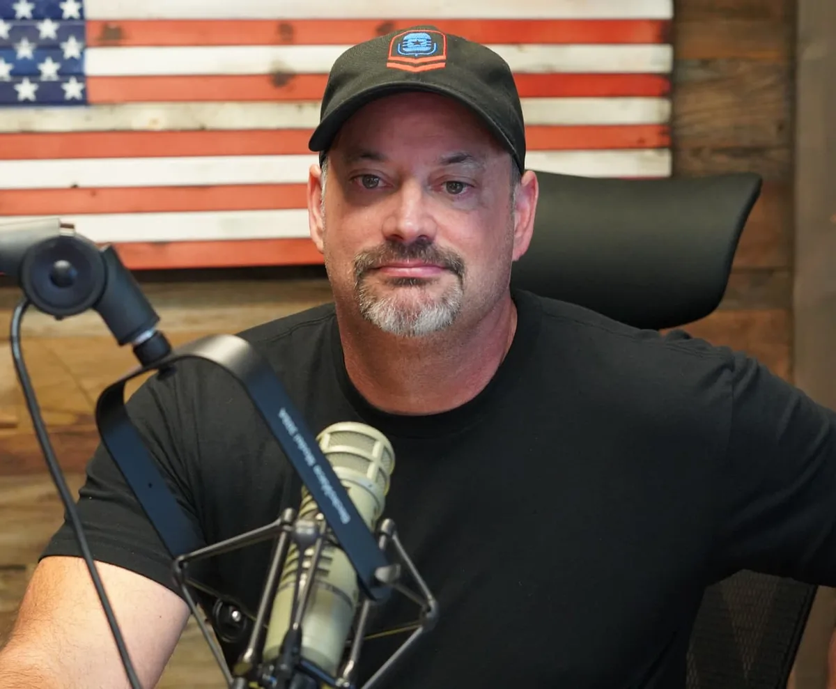 Ed Parcaut wearing a black cap and T-shirt at the studio microphone in front of a wooden American flag